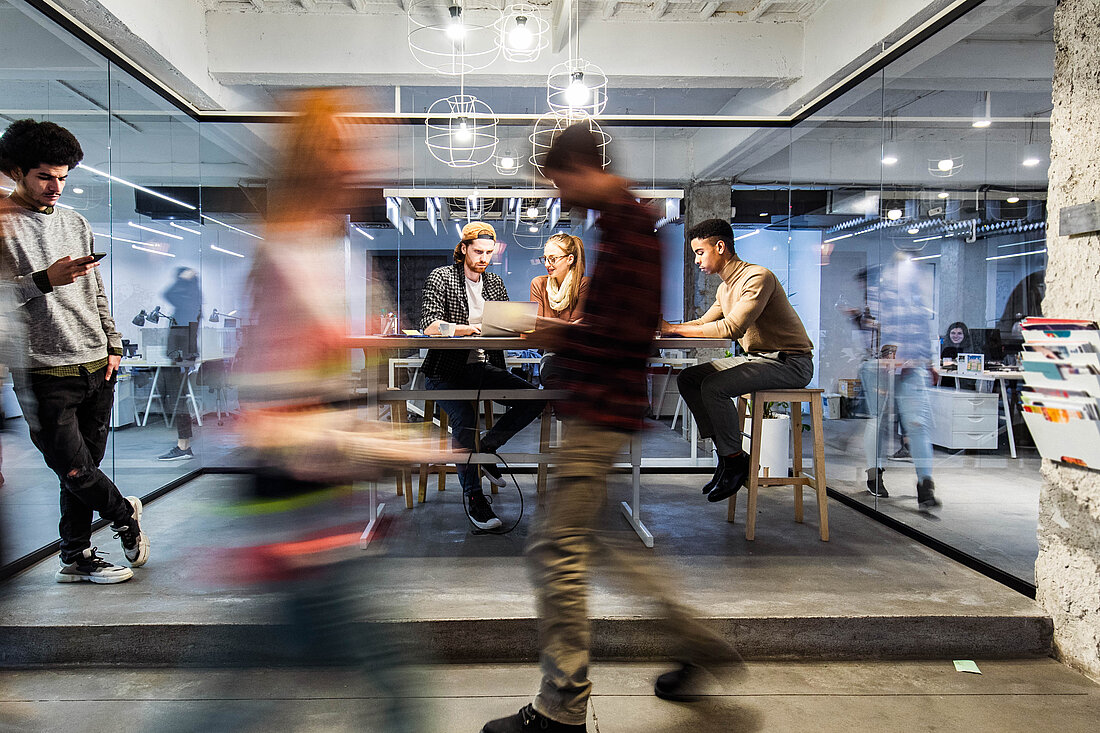 Young people work and talk around a standing table in a startup coworking office, while others move through the workspace.