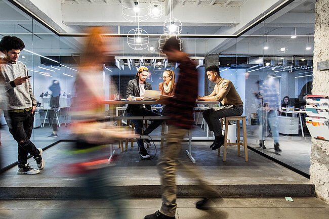 Young people work and talk around a standing table in a startup coworking office, while others move through the workspace.