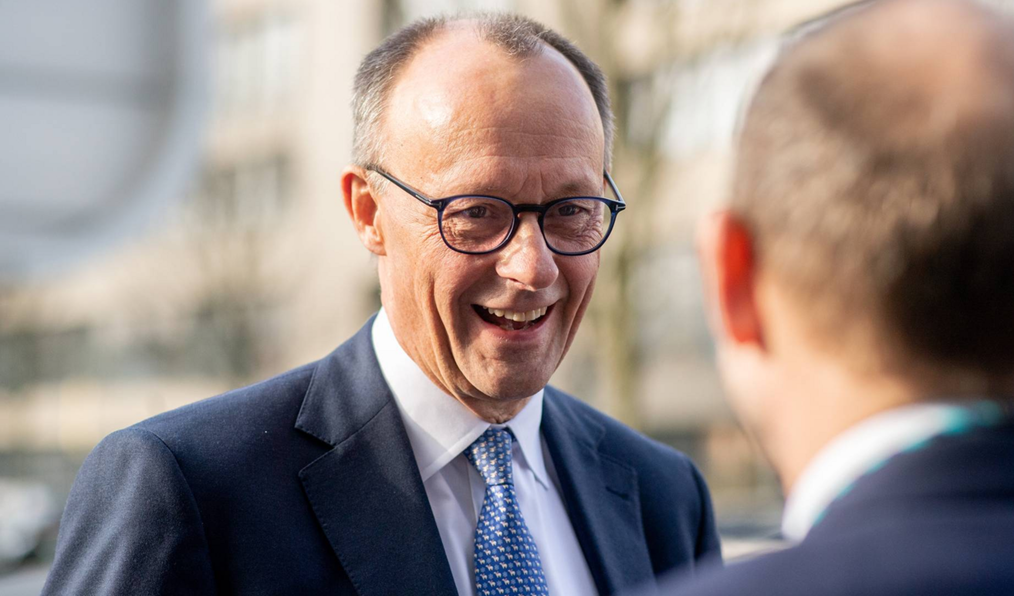 Friedrich Merz, frontally depicted, laughing in a dark suit with glasses.