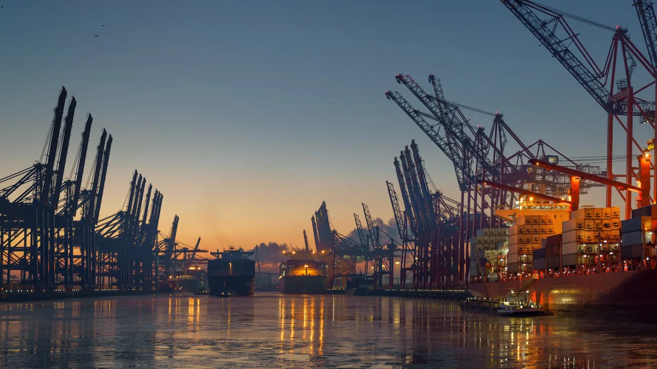 Night harbor view with cranes, container ships, and reflections on the water.